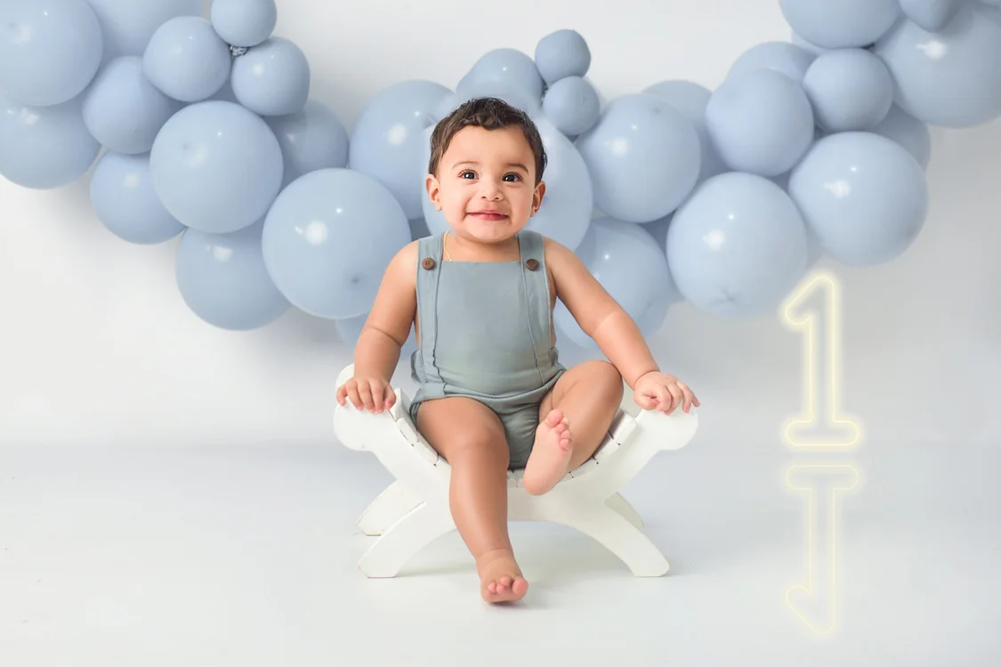 Bebé sentado en una banqueta blanca delante de globos celestes en su sesión de primer añito
