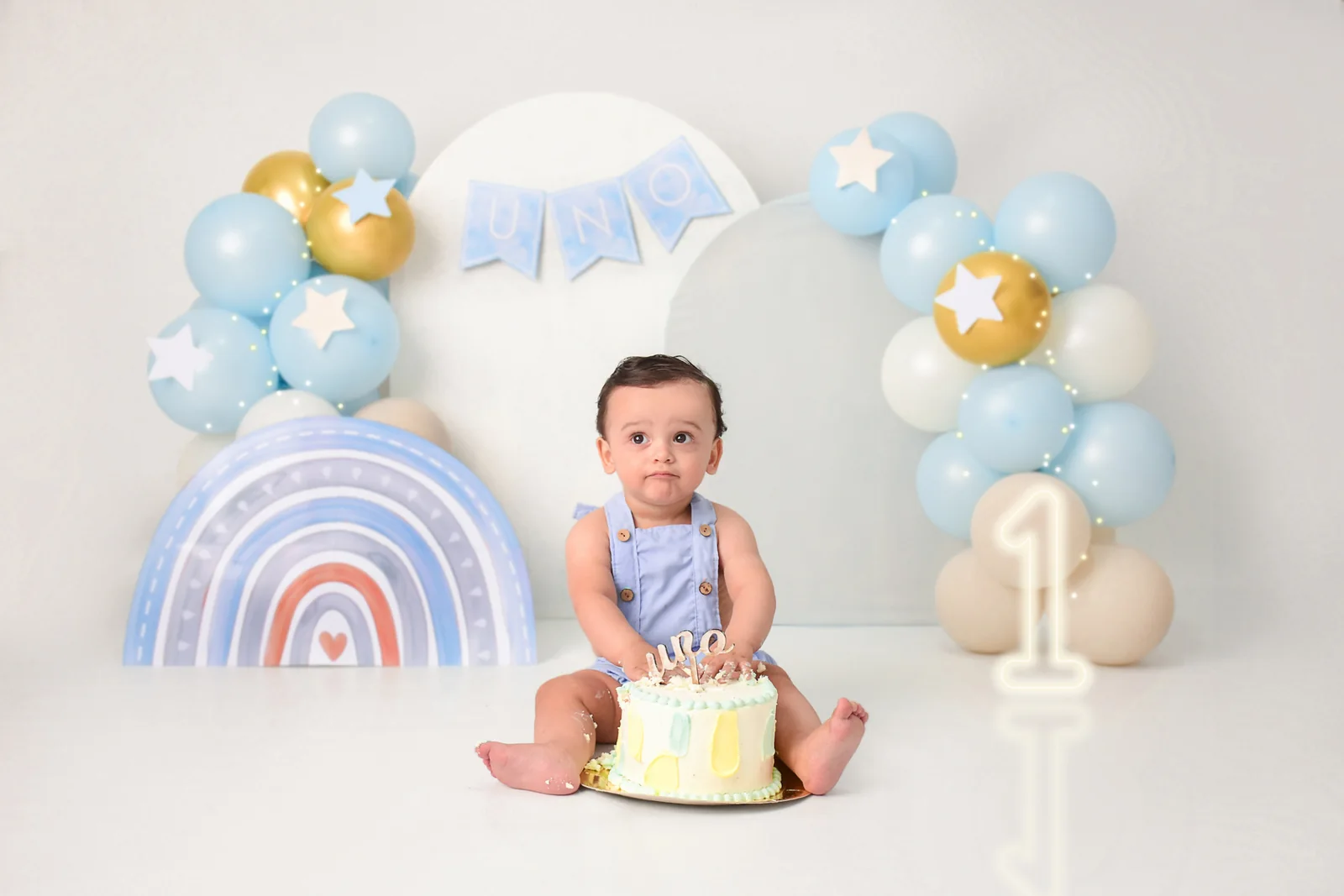 Bebé con torta amarilla delante de un arcoíris de globos