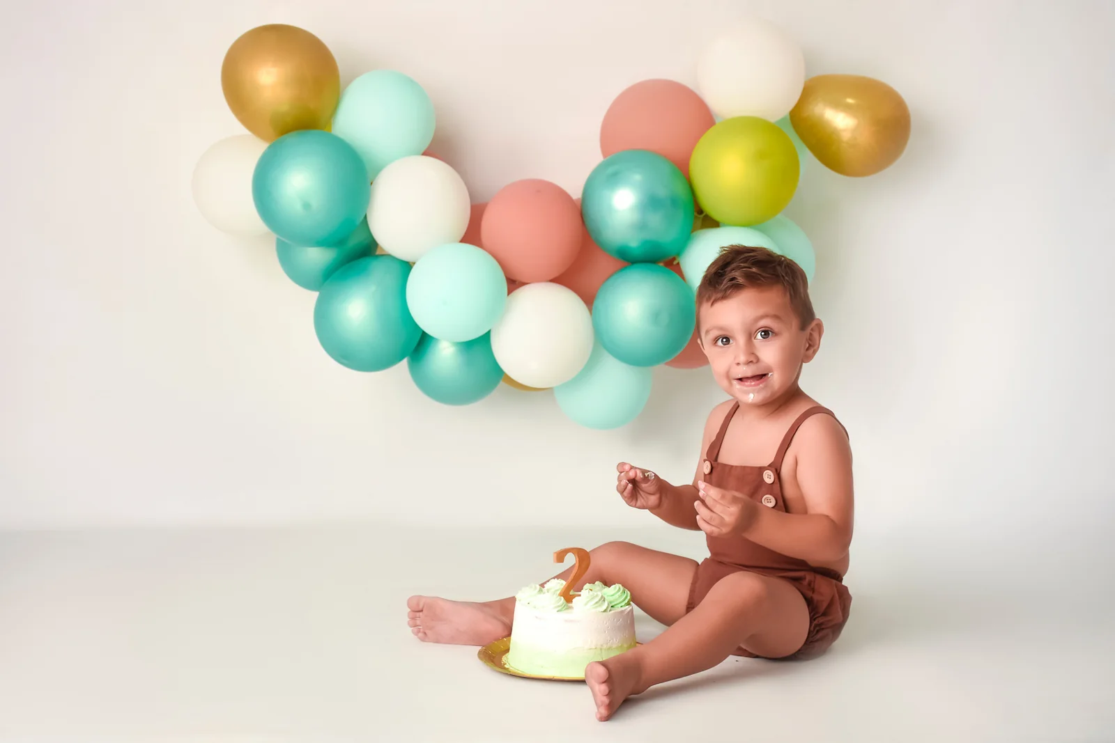 Nene descalzo junto a su torta con globos verdes y dorados