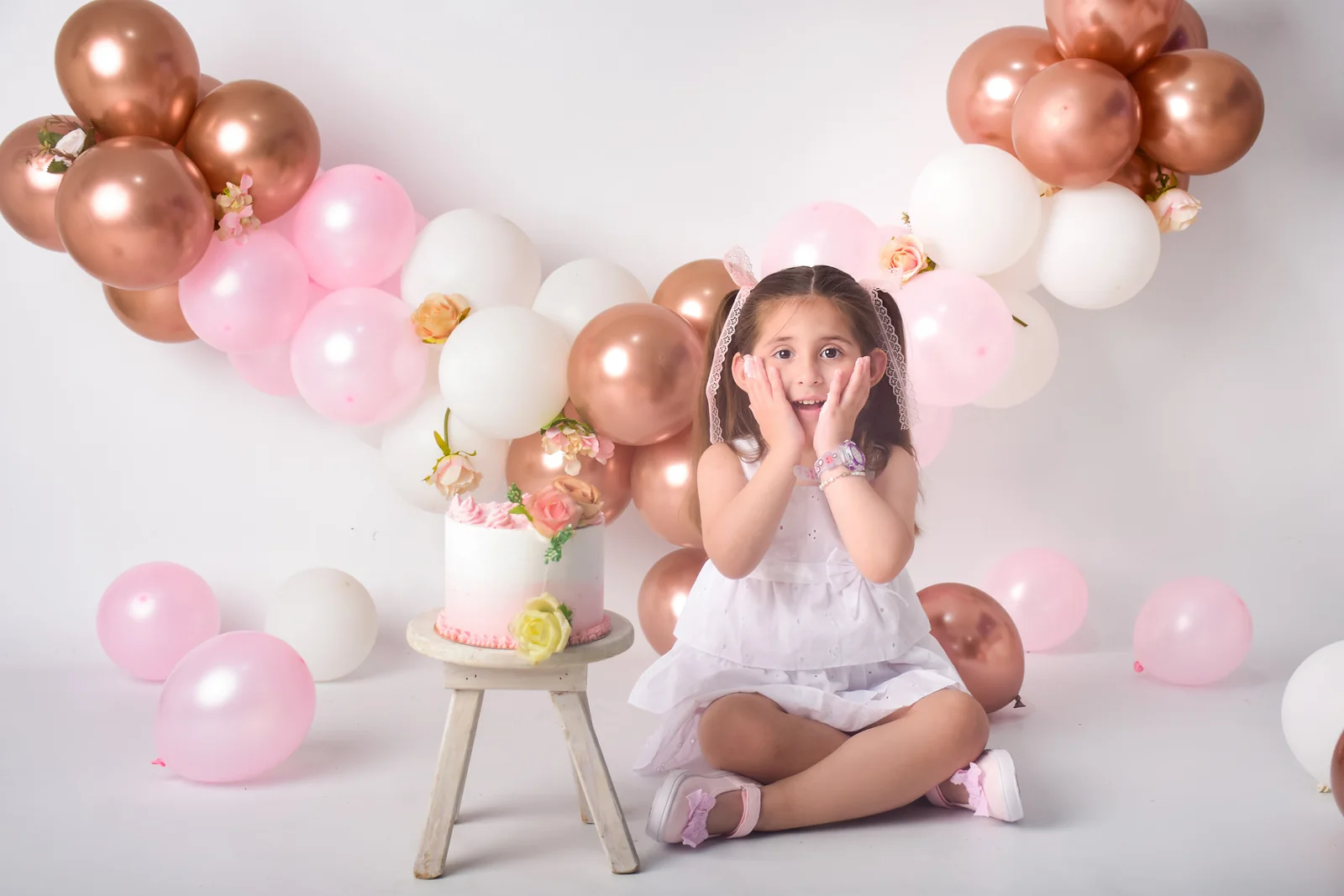 Nena con vestido blanco junto a su torta y globos color cobre